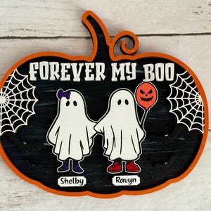 May include: A Halloween-themed wooden pumpkin decoration with the text "FOREVER MY BOO." It features two cartoon ghosts holding hands, one with a purple bow and the other with a balloon. The names "Shelby" and "Ravyn" are below the ghosts.
