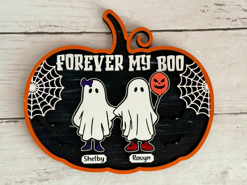 May include: A Halloween-themed wooden pumpkin decoration with the text "FOREVER MY BOO." It features two cartoon ghosts holding hands, one with a purple bow and the other with a balloon. The names "Shelby" and "Ravyn" are below the ghosts.