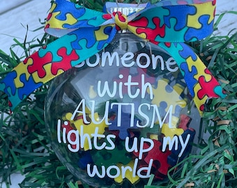 Puede incluir: Un adorno de Navidad de vidrio transparente con una cinta blanca y un estampado de piezas de rompecabezas. El adorno tiene el texto "Someone with AUTISM lights up my world" en el interior.