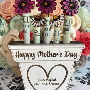May include: Wooden planter with the words "Happy Mother's Day" and "From Crystal, Alex, and Brecken" engraved on it. Three pink flowers are attached to rolled up dollar bills.