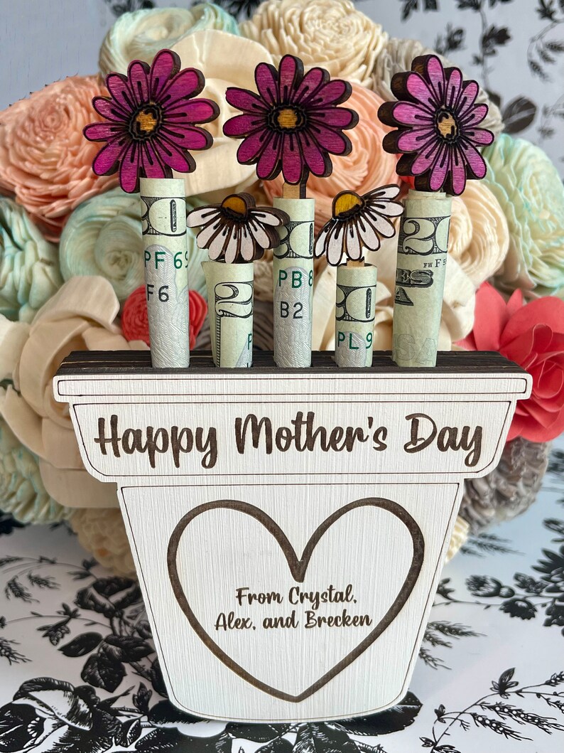 May include: Wooden planter with the words "Happy Mother's Day" and "From Crystal, Alex, and Brecken" engraved on it. Three pink flowers are attached to rolled up dollar bills.