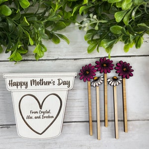 May include: Wooden craft with the text "Happy Mother's Day" and a heart with the text "From Crystal, Alex, and Brecken". Five wooden flower picks with pink and white flowers.
