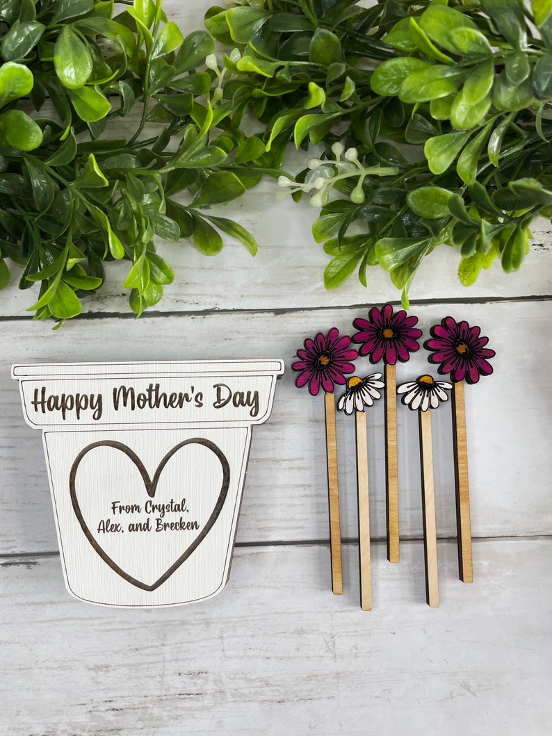 May include: Wooden craft with the text "Happy Mother's Day" and a heart with the text "From Crystal, Alex, and Brecken". Five wooden flower picks with pink and white flowers.
