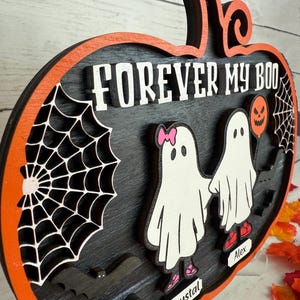 May include: A layered wooden Halloween decoration in the shape of a pumpkin. The design features the text "FOREVER MY BOO", two ghost figures, a spider web, and a bat. The pumpkin has an orange border and a black interior.
