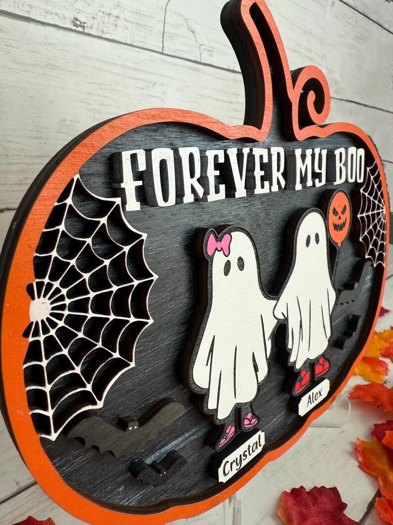 May include: A layered wooden Halloween decoration in the shape of a pumpkin. The design features the text "FOREVER MY BOO", two ghost figures, a spider web, and a bat. The pumpkin has an orange border and a black interior.