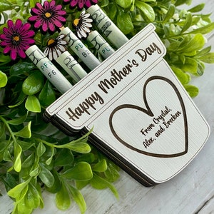 May include: A wooden planter with the text "Happy Mother's Day" and a heart with the text "From Crystal, Alex, and Brecken".  The planter has rolled up dollar bills with flowers attached to the top.