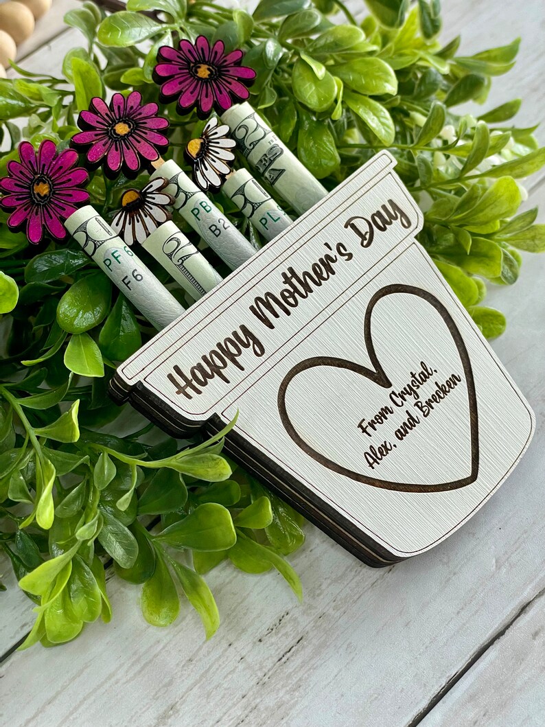 May include: A wooden planter with the text "Happy Mother's Day" and a heart with the text "From Crystal, Alex, and Brecken".  The planter has rolled up dollar bills with flowers attached to the top.