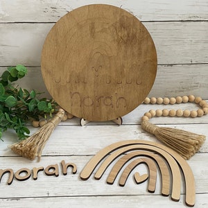 May include: Wooden nursery decor with the name "Norah" engraved on a round plaque and a rainbow with a heart cutout. The name "Norah" is also spelled out in individual wooden letters.