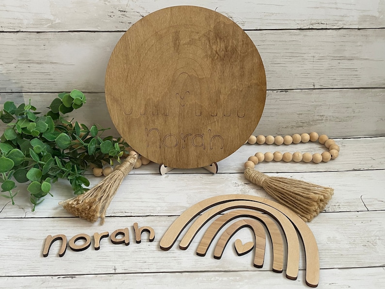 May include: Wooden nursery decor with the name "Norah" engraved on a round plaque and a rainbow with a heart cutout. The name "Norah" is also spelled out in individual wooden letters.