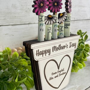May include: Wooden planter with a heart-shaped cutout and the text "Happy Mother's Day" and "From Crystal, Alex, and Brecken". The planter has four pink flowers with white daisy flowers in the center. The flowers are attached to rolled up dollar bills.