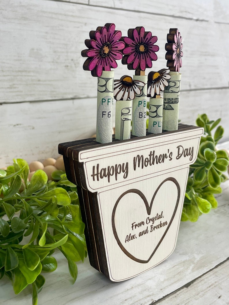 May include: Wooden planter with a heart-shaped cutout and the text "Happy Mother's Day" and "From Crystal, Alex, and Brecken". The planter has four pink flowers with white daisy flowers in the center. The flowers are attached to rolled up dollar bills.