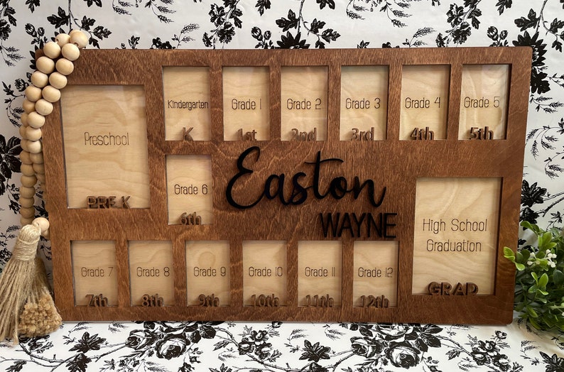Puede incluir: Un marco de fotos de madera con 14 ranuras para fotos, etiquetadas con los grados desde preescolar hasta la graduaci&oacute;n de la escuela secundaria. El marco es marr&oacute;n con un acabado de madera clara y tiene el nombre Easton WAYNE grabado en &eacute;l.