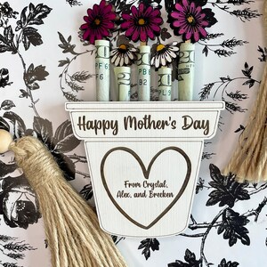 May include: A wooden planter with the words "Happy Mother's Day" and "From Crystal, Alex, and Brecken" engraved on it. The planter has a heart shape cut out and is decorated with rolled up dollar bills and pink and white paper flowers.