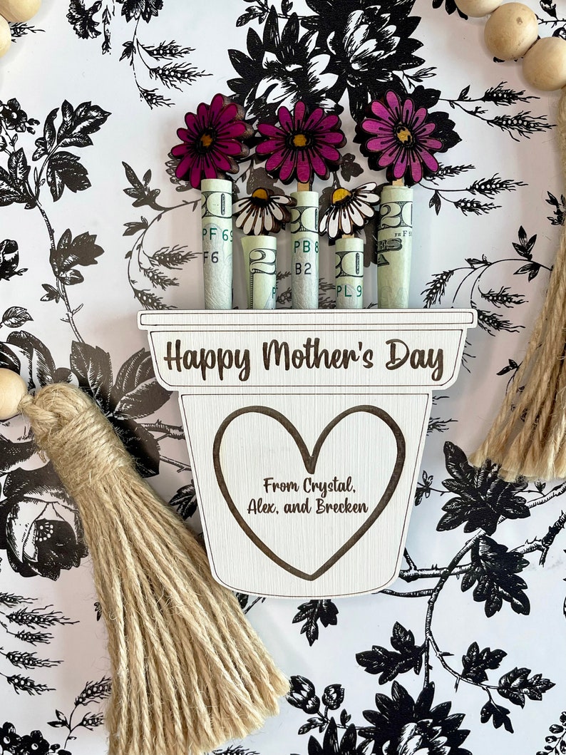 May include: A wooden planter with the words "Happy Mother's Day" and "From Crystal, Alex, and Brecken" engraved on it. The planter has a heart shape cut out and is decorated with rolled up dollar bills and pink and white paper flowers.