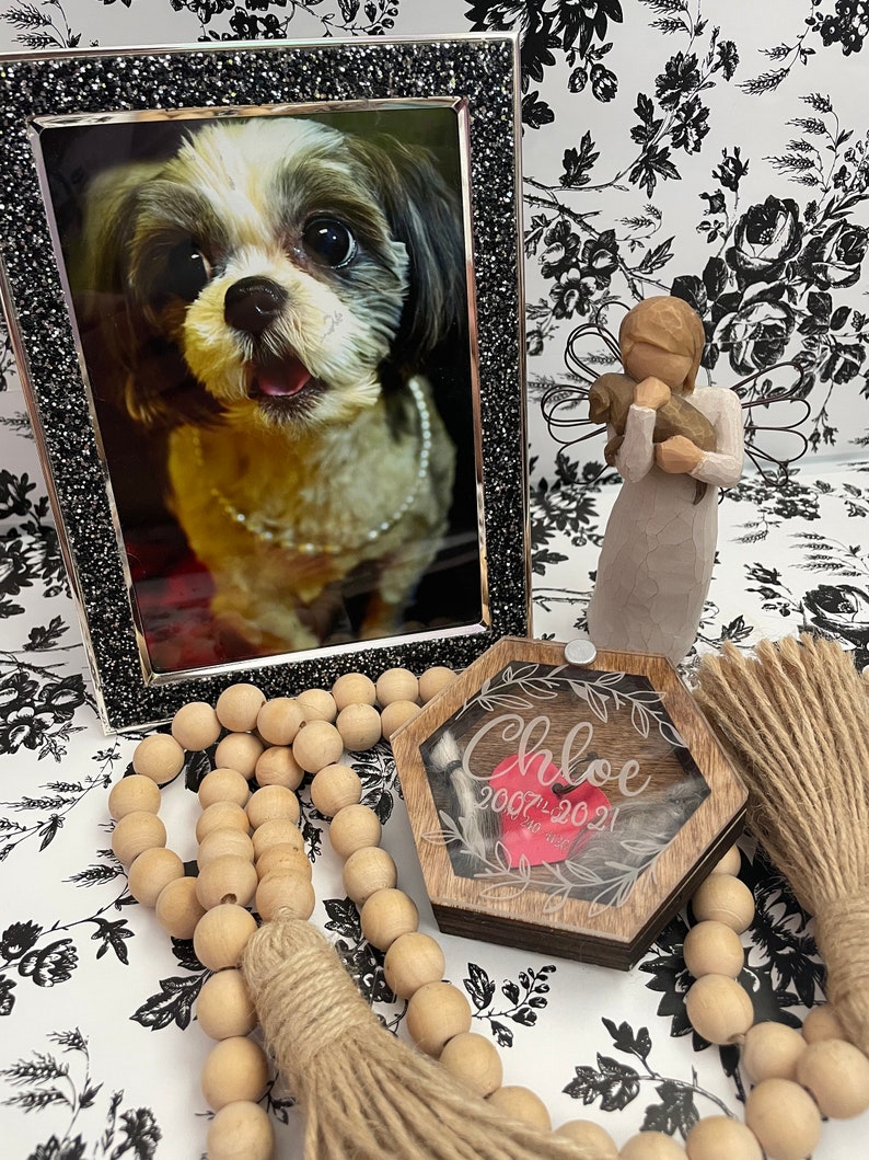 May include: A wooden hexagonal box with a clear lid and a red heart inside. The box has the name "Chloe" engraved on it, along with the dates "2007-2021". The box is surrounded by a wooden bead garland with tassels.