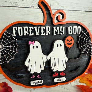 May include: A wooden Halloween decoration in the shape of a pumpkin with the words "FOREVER MY BOO". It features two ghost figures holding hands, a jack-o'-lantern balloon, and spiderwebs. The names "Crystal" and "Alex" are on the bottom.