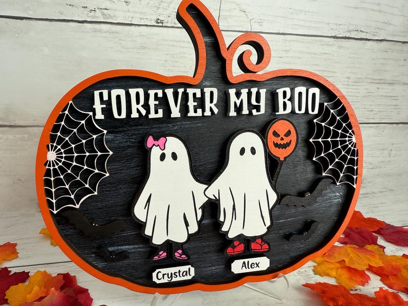 May include: A wooden Halloween decoration in the shape of a pumpkin with the words "FOREVER MY BOO". It features two ghost figures holding hands, a jack-o'-lantern balloon, and spiderwebs. The names "Crystal" and "Alex" are on the bottom.