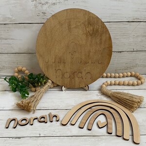 May include: Wooden rainbow wall decor with the name "Norah" and a heart cutout. The set includes a round wooden sign with a rainbow and the name "Norah" and a heart cutout, a set of three wooden rainbow arches, and a wooden name puzzle.