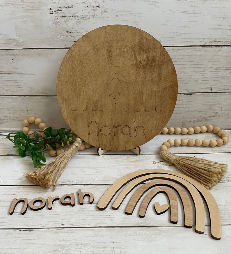 May include: Wooden rainbow wall decor with the name "Norah" and a heart cutout. The set includes a round wooden sign with a rainbow and the name "Norah" and a heart cutout, a set of three wooden rainbow arches, and a wooden name puzzle.