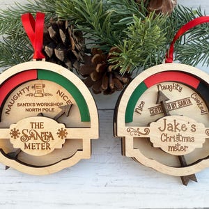 May include: Wooden Christmas ornaments shaped like meters, with red and green sections labeled "Naughty" and "Nice." One reads "The Santa Meter," the other "Jake's Christmas meter." Both have red ribbons.