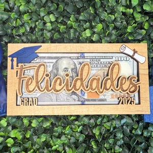 May include: A wooden money holder with a slot for cash. The holder has a graduation cap, diploma, and the word "GRAD" on the front. The text "felicidades 2025" is also on the front of the holder.