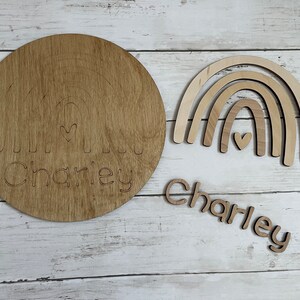 May include: A wooden craft kit for making a rainbow with the name "Charley" engraved on it. The kit includes a round wooden base, a rainbow cutout, and individual letters to spell the name.