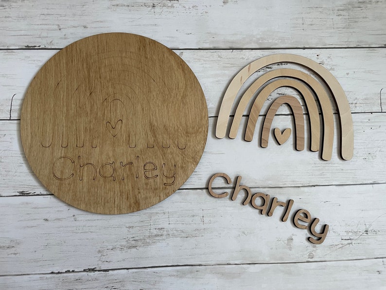 May include: A wooden craft kit for making a rainbow with the name "Charley" engraved on it. The kit includes a round wooden base, a rainbow cutout, and individual letters to spell the name.