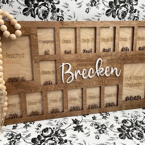 Puede incluir: Un marco de fotos de madera con 14 ranuras para fotos, etiquetadas con los grados desde preescolar hasta la graduaci&oacute;n de la escuela secundaria. El marco est&aacute; personalizado con el nombre "Brecken".