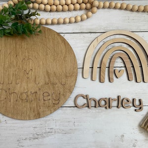 May include: Wooden craft kit with a round wooden plaque, a rainbow cutout, and a separate name cutout. The plaque has a rainbow design with a heart in the center and the name "Charley" written below. The rainbow cutout is made of three layers of wood and has a heart in the center. The name cutout is made of wood and spells out "Charley".