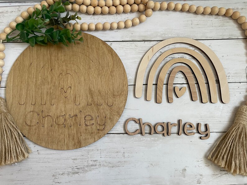 May include: Wooden craft kit with a round wooden plaque, a rainbow cutout, and a separate name cutout. The plaque has a rainbow design with a heart in the center and the name "Charley" written below. The rainbow cutout is made of three layers of wood and has a heart in the center. The name cutout is made of wood and spells out "Charley".