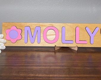 May include: Wooden puzzle with the name "MOLLY" spelled out in pink and purple letters. The puzzle has a pink flower and butterfly on either end.