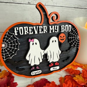 May include: A Halloween-themed wooden sign in the shape of a pumpkin. The sign is black with orange trim and features the text "FOREVER MY BOO" in white. Two ghost figures are holding hands. One ghost has a pink bow and the other holds a pumpkin balloon.