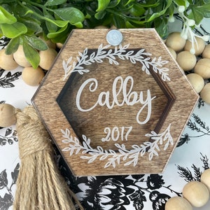 May include: A hexagonal wooden ornament with a clear acrylic insert. The ornament is decorated with a white floral design and the name "Calby" and the year "2017" are inscribed in white. The ornament has a brown jute tassel.