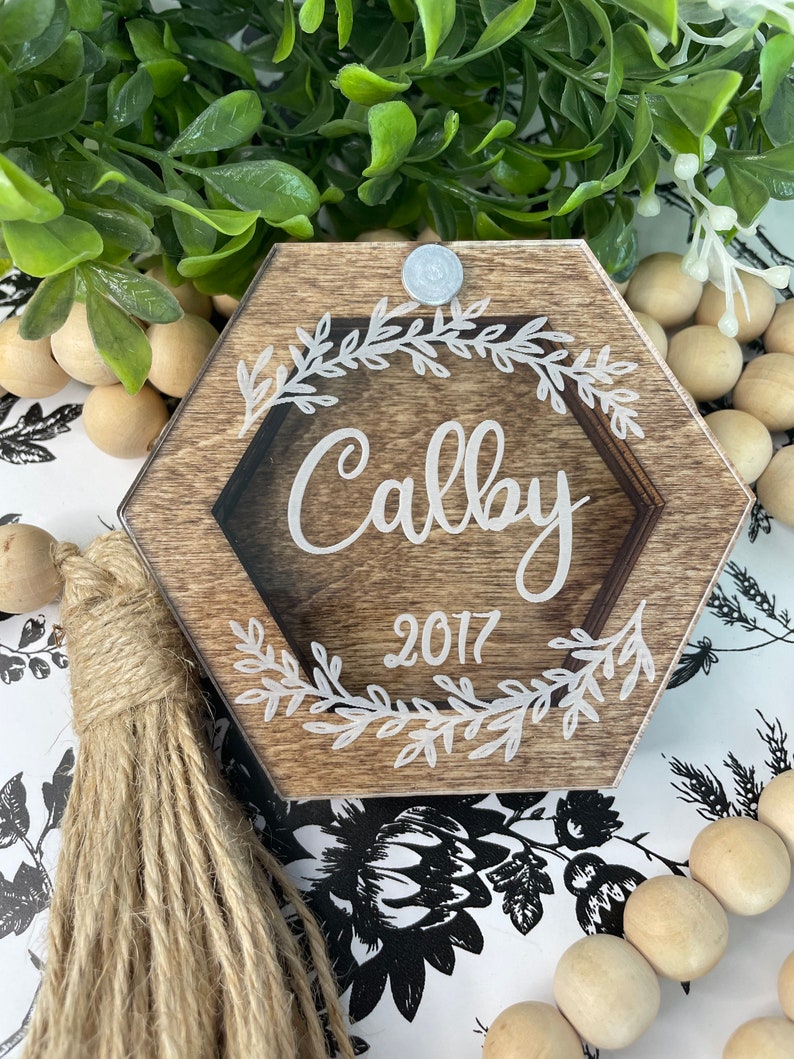 May include: A hexagonal wooden ornament with a clear acrylic insert. The ornament is decorated with a white floral design and the name "Calby" and the year "2017" are inscribed in white. The ornament has a brown jute tassel.