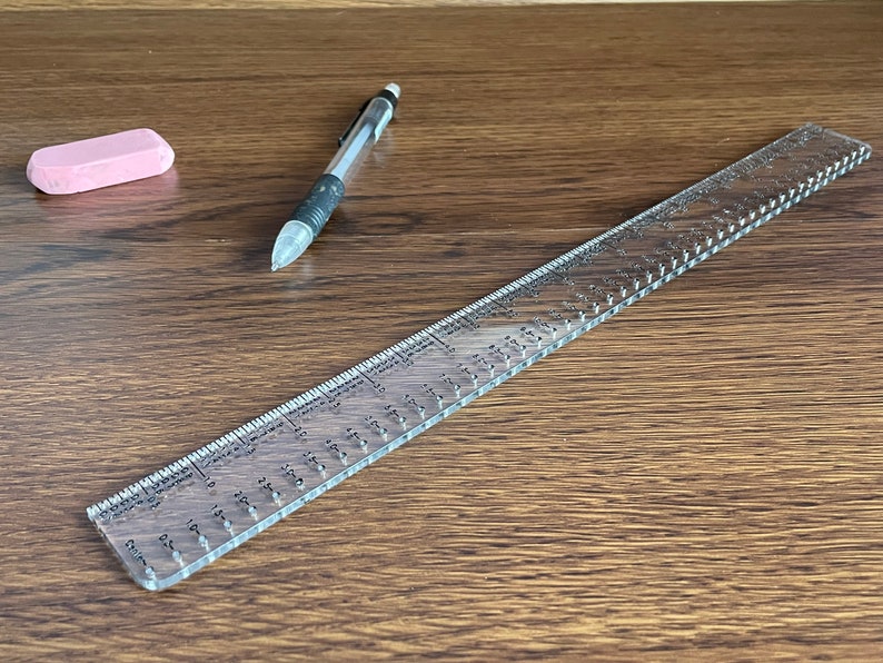 K&ouml;nnte beinhalten: Ein durchsichtiges Lineal aus Kunststoff mit schwarzen Markierungen und einem rosa Radiergummi auf einer Holzoberfl&auml;che.