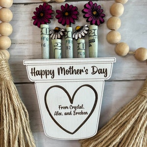 May include: A wooden planter with a heart-shaped cutout and the text "Happy Mother's Day" and "From Crystal, Alex, and Brecken". The planter has five pink flowers with white daisy flowers growing from rolled up dollar bills.