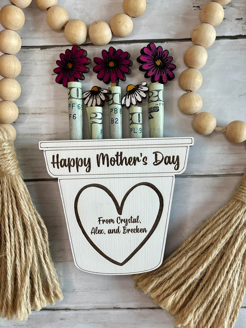 May include: A wooden planter with a heart-shaped cutout and the text "Happy Mother's Day" and "From Crystal, Alex, and Brecken". The planter has five pink flowers with white daisy flowers growing from rolled up dollar bills.