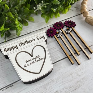 May include: Wooden craft with the text "Happy Mother's Day" and a heart with the text "From Crystal, Alex, and Brecken". The craft is decorated with pink and white flowers.