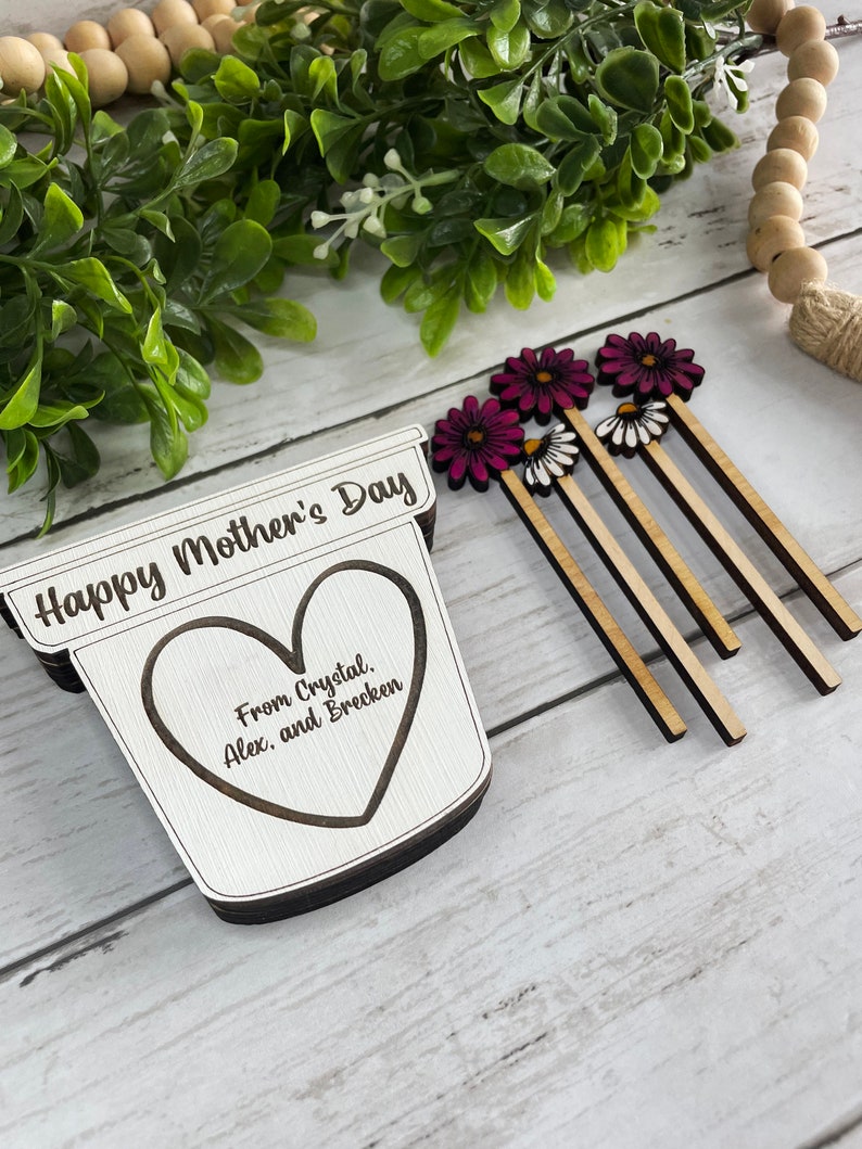 May include: Wooden craft with the text "Happy Mother's Day" and a heart with the text "From Crystal, Alex, and Brecken". The craft is decorated with pink and white flowers.