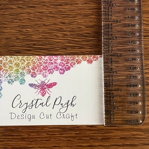 K&ouml;nnte beinhalten: Eine wei&szlig;e Visitenkarte mit einem Regenbogen-Glitzer-Wabenmuster und einer Bienenillustration. Der Text auf der Karte lautet "Crystal Pugh Design Cut Craft".