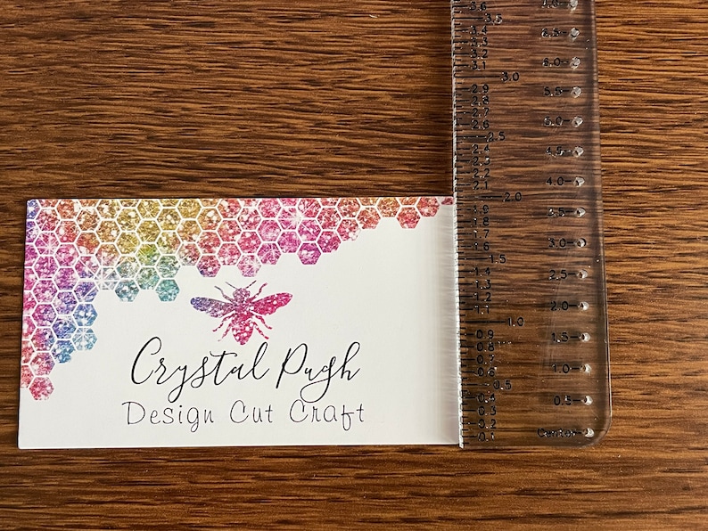 K&ouml;nnte beinhalten: Eine wei&szlig;e Visitenkarte mit einem Regenbogen-Glitzer-Wabenmuster und einer Bienenillustration. Der Text auf der Karte lautet "Crystal Pugh Design Cut Craft".