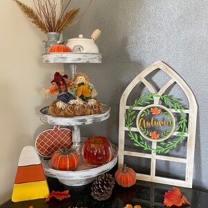 May include: A three-tiered tray with fall-themed decorations. Includes pumpkins, scarecrows, and a decorative window with the word "Autumn". The tray is white with a distressed finish, and the items are in shades of orange, brown, and yellow.