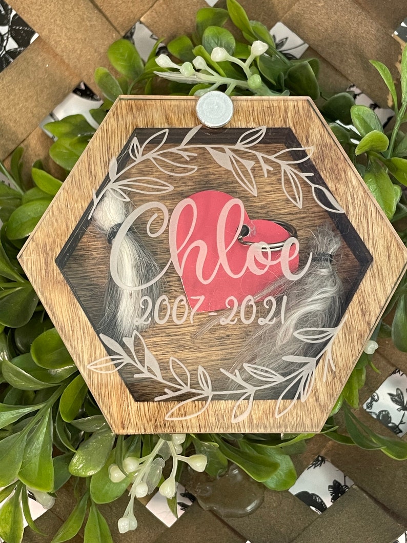 May include: A wooden hexagonal box with a glass insert. The box has a white inscription that reads "Chloe 2007-2021" and a red heart inside. The box is surrounded by a leafy design.