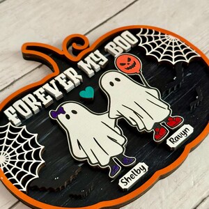 May include: A Halloween-themed wooden pumpkin decoration with the words "FOREVER MY BOO" in white. Features two ghost figures, a heart, and spiderwebs. The names "Shelby" and "Ravyn" are included. The pumpkin has an orange border and black interior.