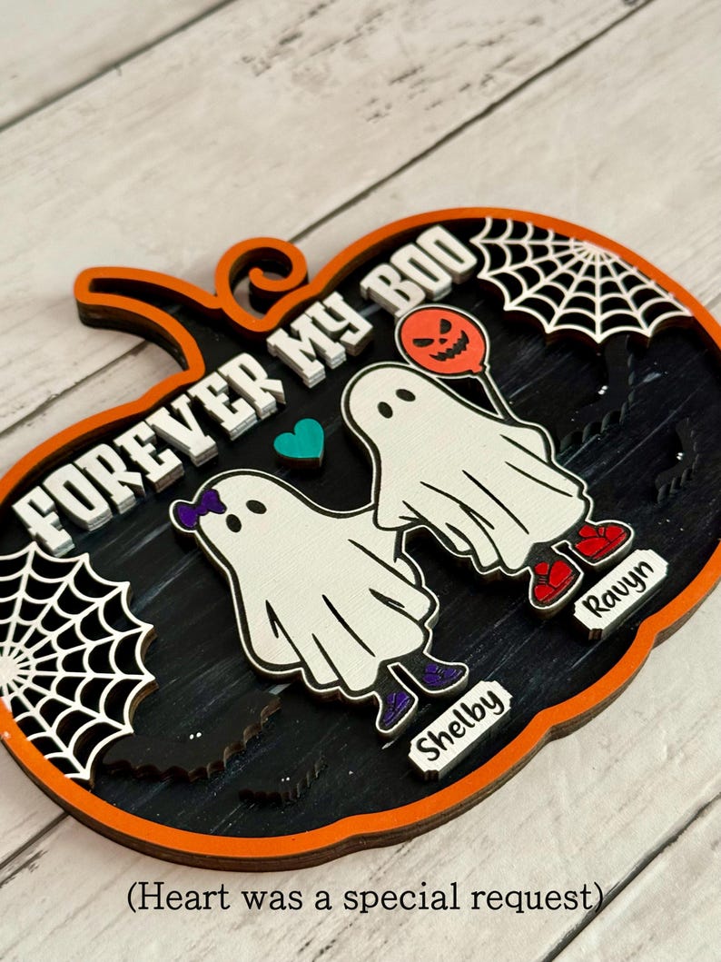 May include: A Halloween-themed wooden pumpkin decoration with the words "FOREVER MY BOO" in white. Features two ghost figures, a heart, and spiderwebs. The names "Shelby" and "Ravyn" are included. The pumpkin has an orange border and black interior.