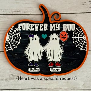 May include: A Halloween-themed pumpkin-shaped wooden sign with the text "FOREVER MY BOO". It features two ghost figures holding hands, with the names "Shelby" and "Ravyn" below. One ghost has a purple bow and the other holds a jack-o'-lantern balloon.