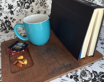 May include: A wooden tray with a personalized inscription "Heather" and a floral design. The tray has a built-in cup holder, a small compartment for snacks, and a book holder. The tray is brown and has a natural wood finish.