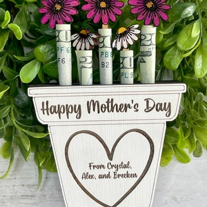 May include: A wooden planter with the text "Happy Mother's Day" and a heart with the text "From Crystal, Alex, and Brecken".  The planter has four rolled up dollar bills with pink and white flowers on top.