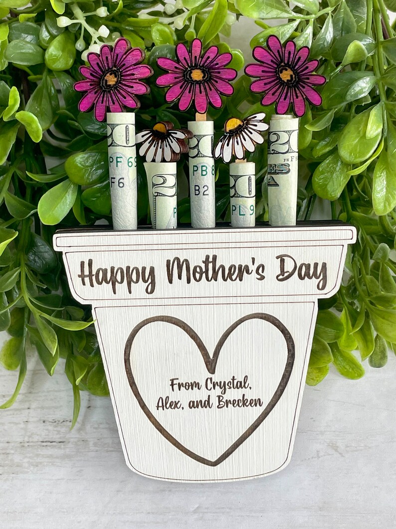 May include: A wooden planter with the text "Happy Mother's Day" and a heart with the text "From Crystal, Alex, and Brecken".  The planter has four rolled up dollar bills with pink and white flowers on top.