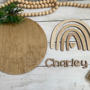 May include: Wooden rainbow wall decor with a heart in the center and the name Charley written in cursive on a separate piece of wood. The name Charley is also written on a round wooden plaque with a rainbow design.